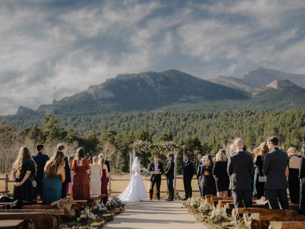 a wedding ceremony takes place in the meadow at wild basin lodge outside of Rocky Mountain national park with the mountains in the background