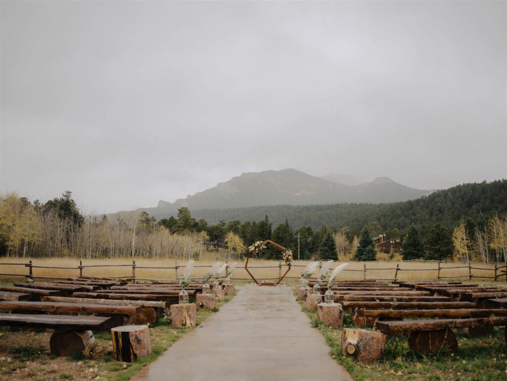 a ceremony site full of wooden benches and a beautiful wooden arch sit at the base of a mountain on a foggy day at wild basin lodge before a wedding ceremony