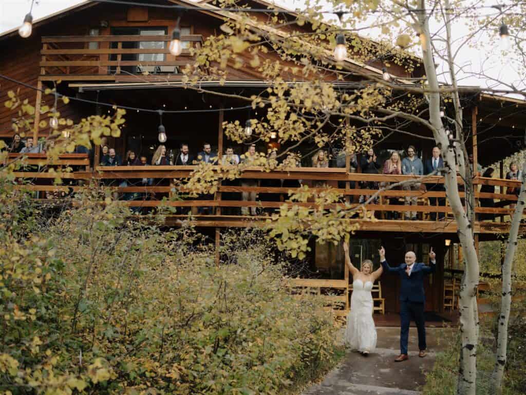 bride and groom enter their reception at wild basin lodge as they run into the amphitheater area with the lodge behind them as their friends and family watch