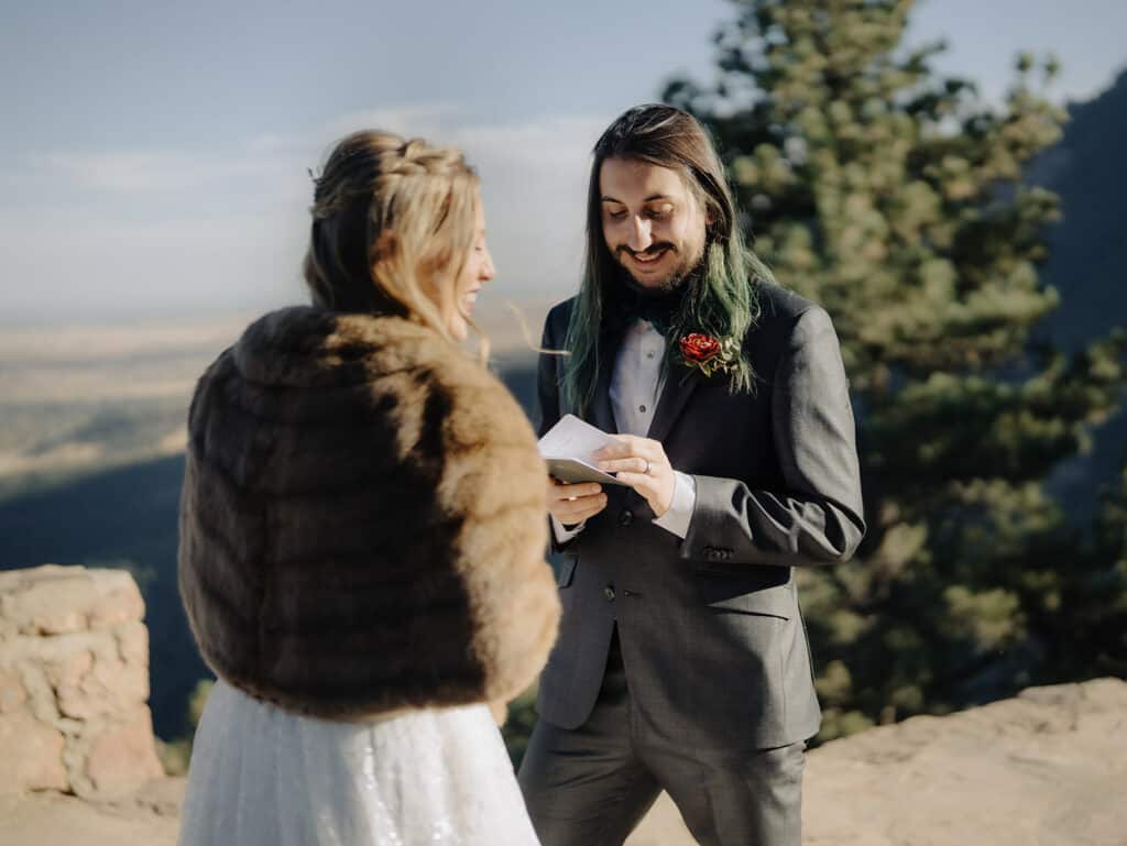 a groom reads his vows to the bride in a white dress wearing fur as they stand at sunrise amphitheater overlooking boulder for their wedding ceremony