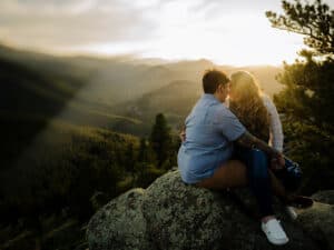 lesbian couple sits on the edge of a mountain at sunset nuzzling into each other for their boulder engagement photographs by Alex medvick