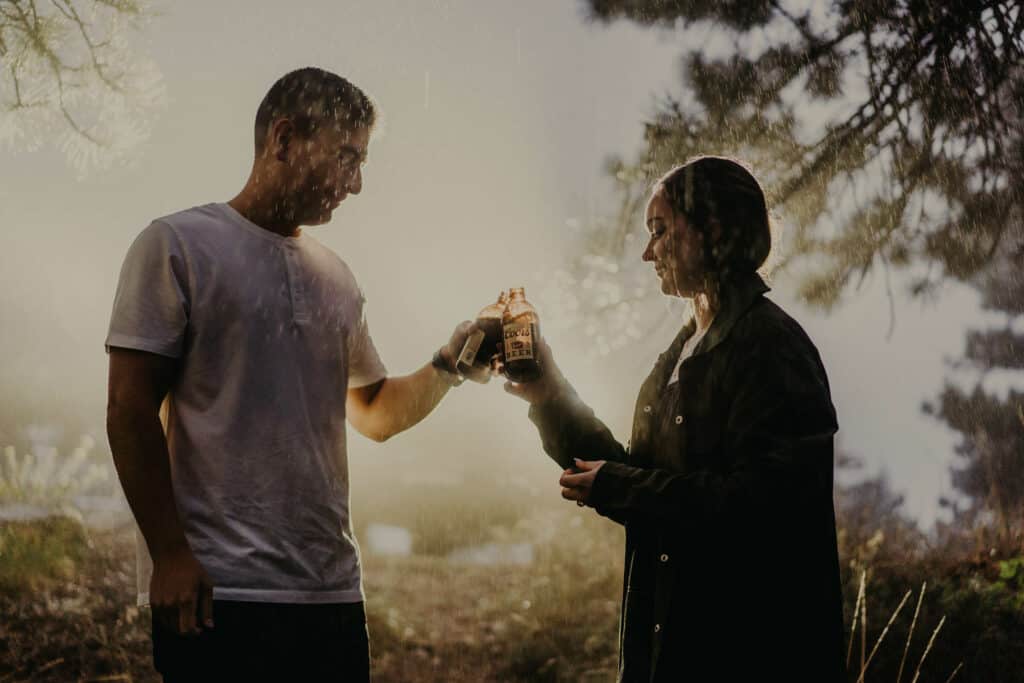a couple toasts with coors light in the rain in front of their car as the headlights illuminate them from behind for their boulder engagement session photographed by Alex medvick