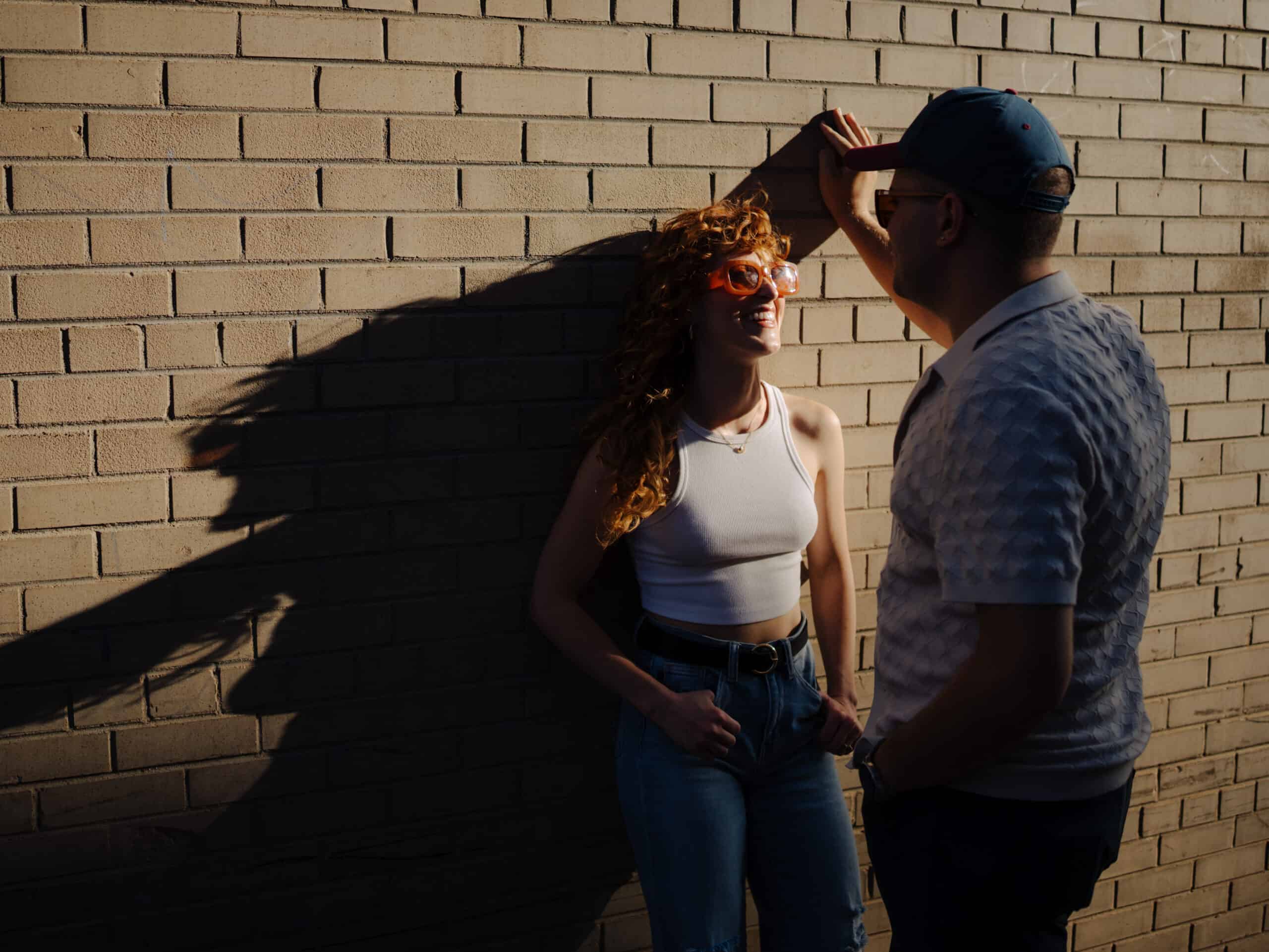 man leans against the wall in super cool sunset light while his fiance looks lovingly at him in a cool alley in boulder colorado for their engagement session with Alex medvick